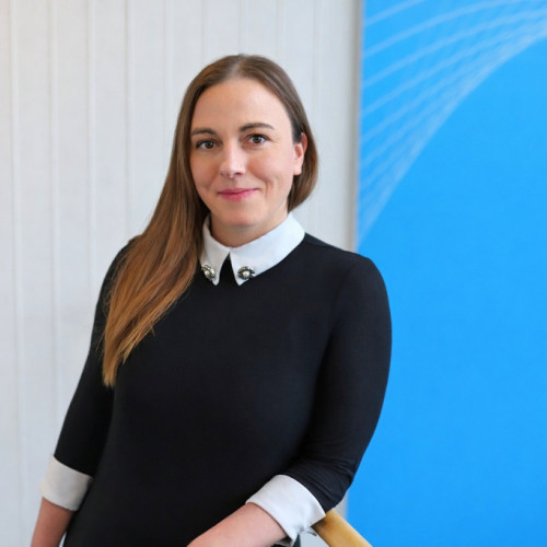 Portrait of a woman with long brown hair wearing a black top with a white collar, smiling slightly in front of a modern office background with a blue banner.